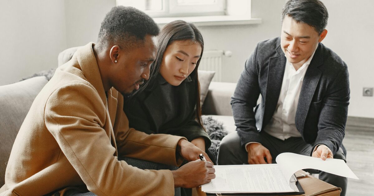 A Black man, an Asian woman, and an Asian man sitting around a table, looking at a document that the Black man is signing.