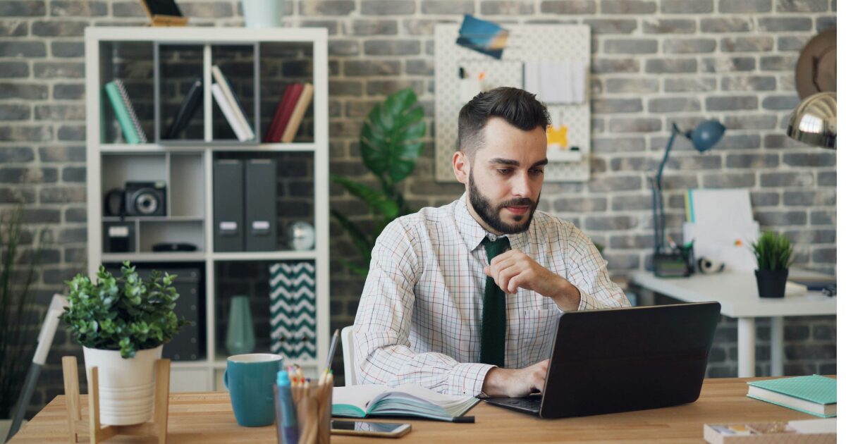 A Caucasian man typing on his laptop, sitting at a desk wearing a white button-up shirt and black tie.