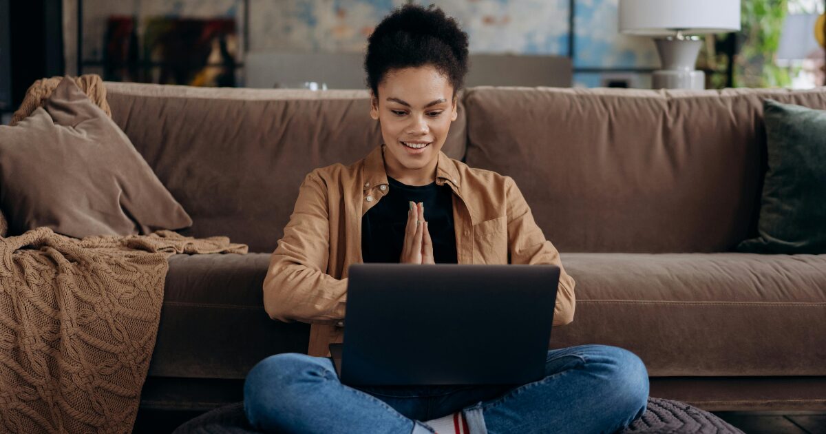 Young Black woman sitting cross-legged, working on her laptop, in front of a brown couch.