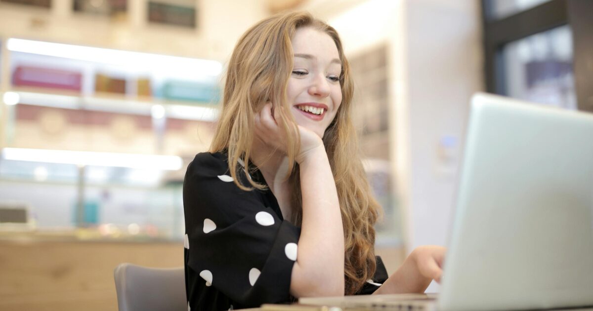 Young Caucasian woman smiling while reading on her laptop, in a black polka dot shirt.