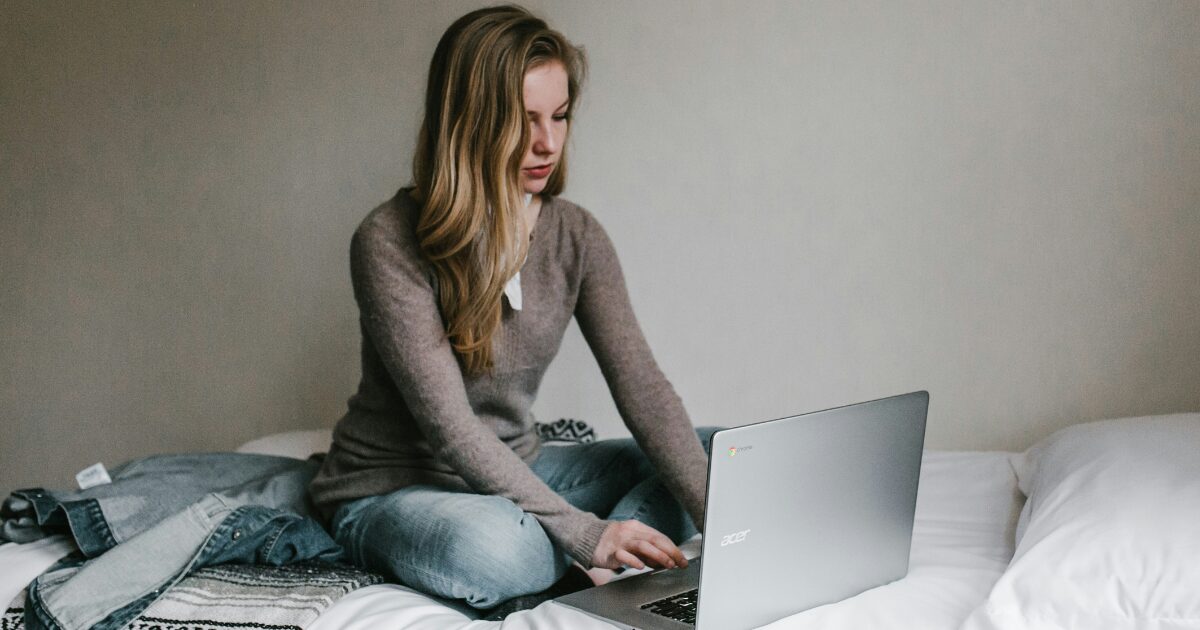 Young Caucasian woman sitting on her bed typing on a laptop. 