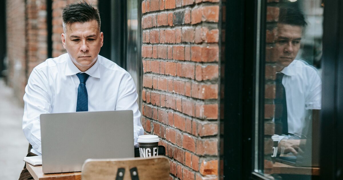 A middle-aged Caucasian man typing on his laptop, sitting on a cafe patio in a white button-up shirt and dark blue tie.