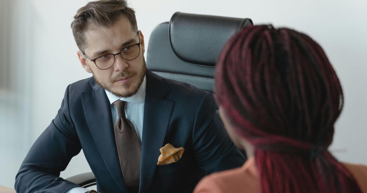 A Caucasian man in a suit talking to a Black woman, her back is facing us