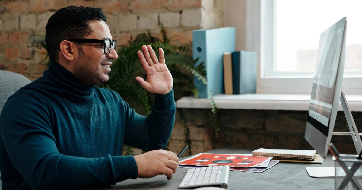 Middle-Eastern middle-aged man wearing a turquoise turtle neck, saying hi on a video call on his computer