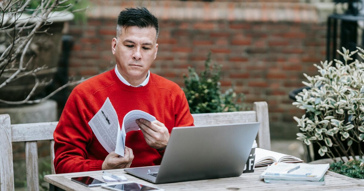 Middle-aged Caucasian man wearing an orange sweater, sitting at a desk on a patio looking over paperwork