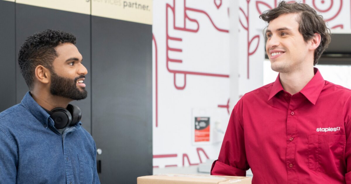 Your male Staples associate helping a young male East Indian customer. The associate is wearing a red button-up Staples shirt and the customer is wearing a denim button-up shirt.