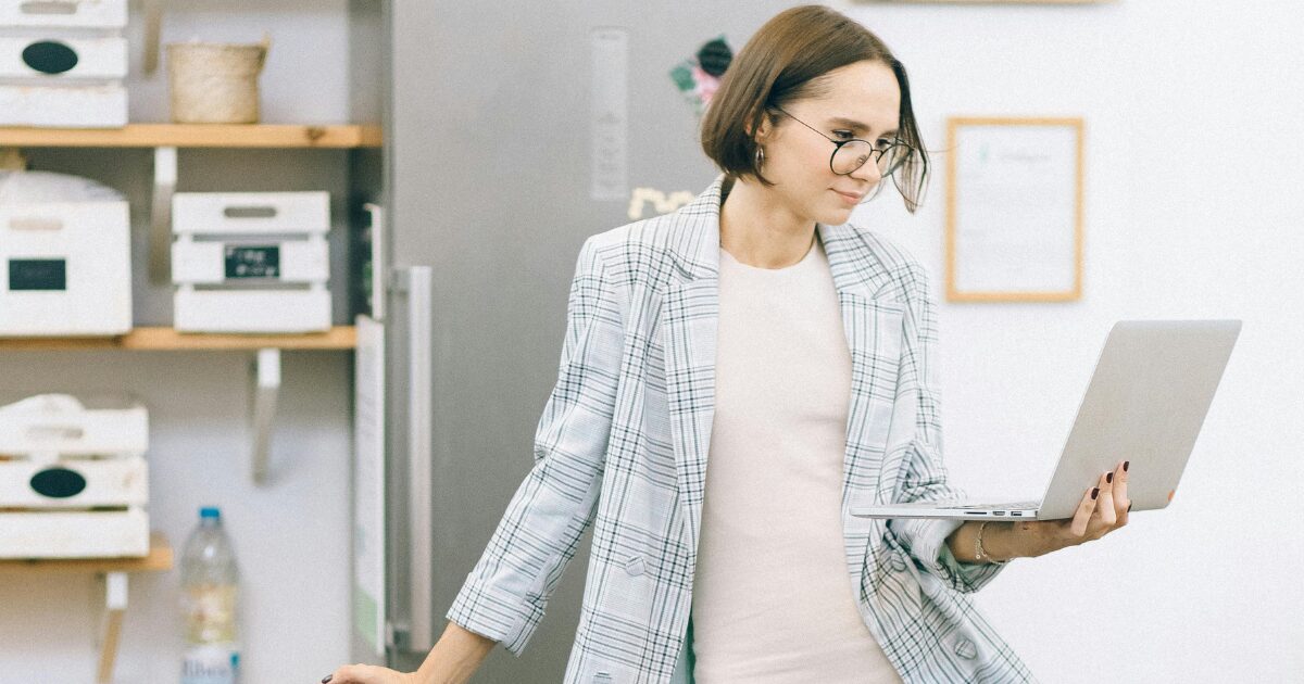 A young Caucasian woman in a plaid blazer, reading something on her laptop while standing, with her laptop in her hand.