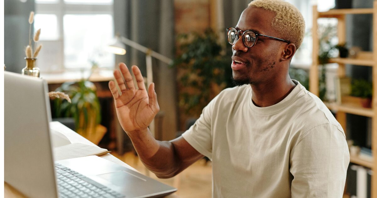 A young Black woman man on a video call, sitting at his desk, waving to the camera.