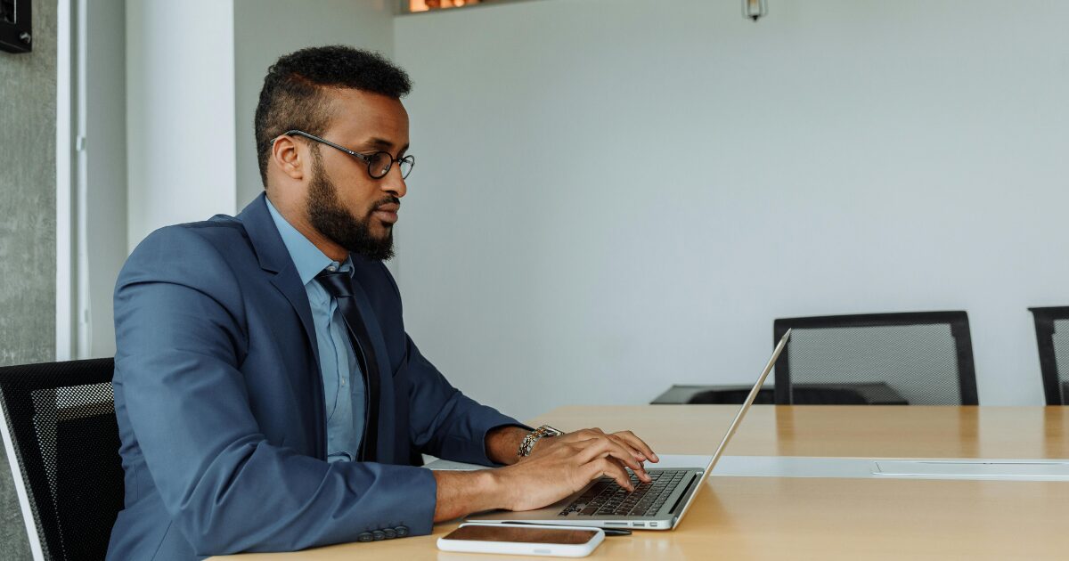 An adult South Asian man sitting at a wooden desk, typing on a laptop. He is wearing a blue blazer and dress shirt. 