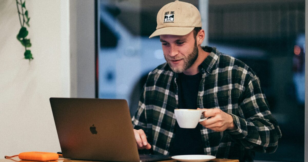 Young Caucasian man in a plaid button-up shirt, sitting at a table drinking a cup of coffee, reading on his laptop.