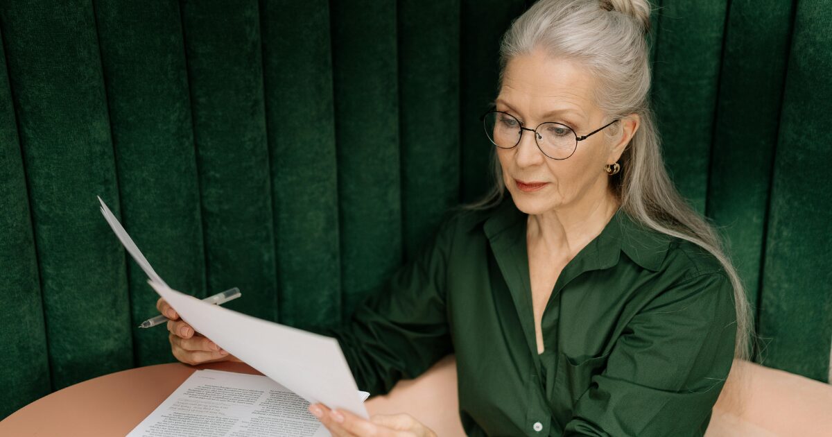 Senior Caucasian woman sitting at a table looking over paperwork. She is wearing a green button-up shirt, red lipstick, and glasses.