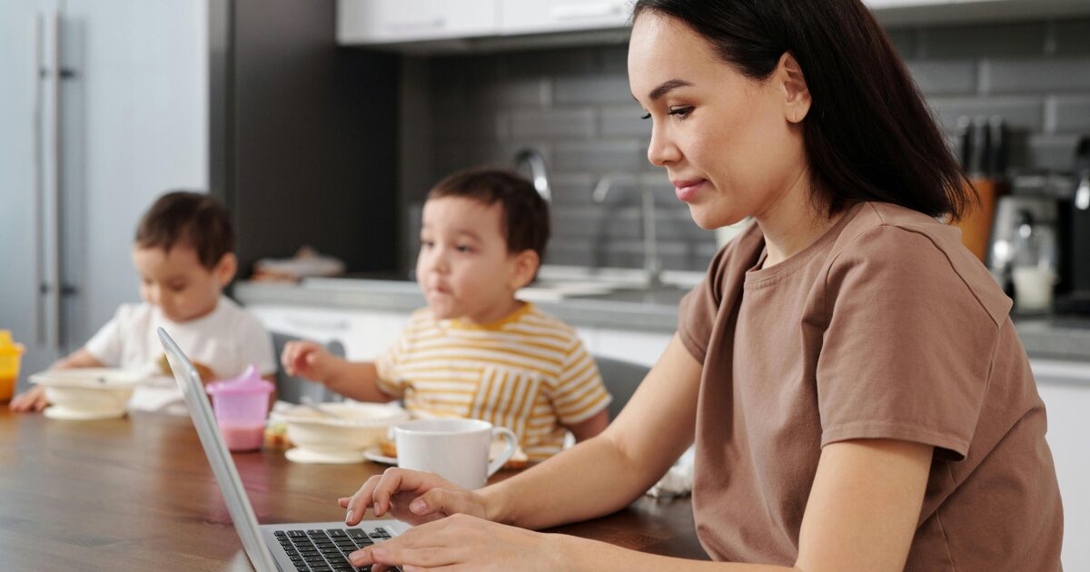 East Asian woman writing on her laptop, sitting at a kitchen table with two young children. 