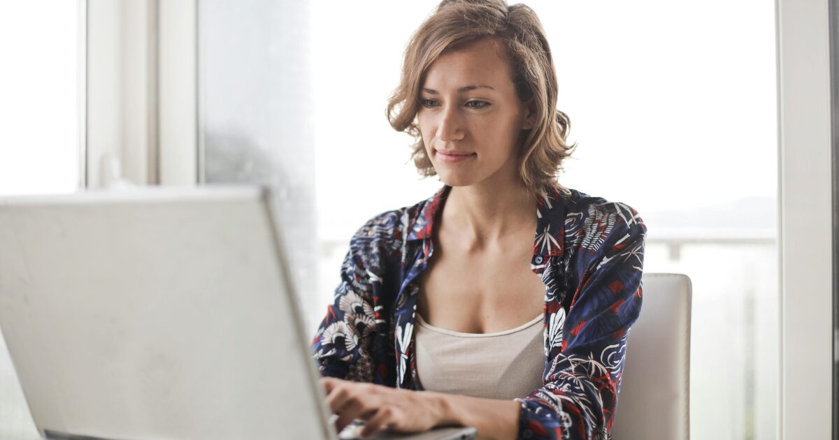 Caucasian woman with short blonde hair creating a document on her laptop, sitting at a table. 