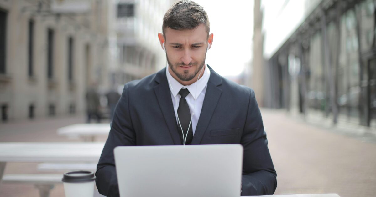 Caucasian man in a suit, typing on a laptop sitting at a patio table outside.