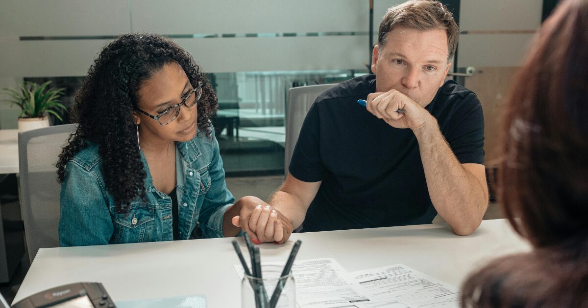 A Black woman holding hands with a Caucasian man, sitting at a table with paperwork in front of them. She is wearing a jean jacket and he is wearing a black t-shirt.