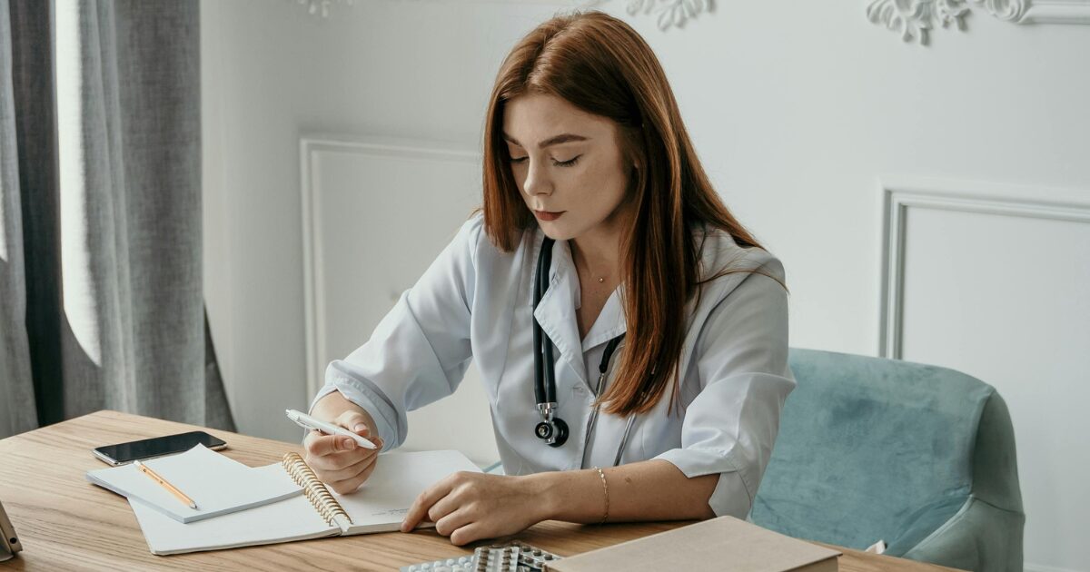 A Young Cacuasian woman wearing a doctor's white coat with a stethoscope on. She is signing paperwork.