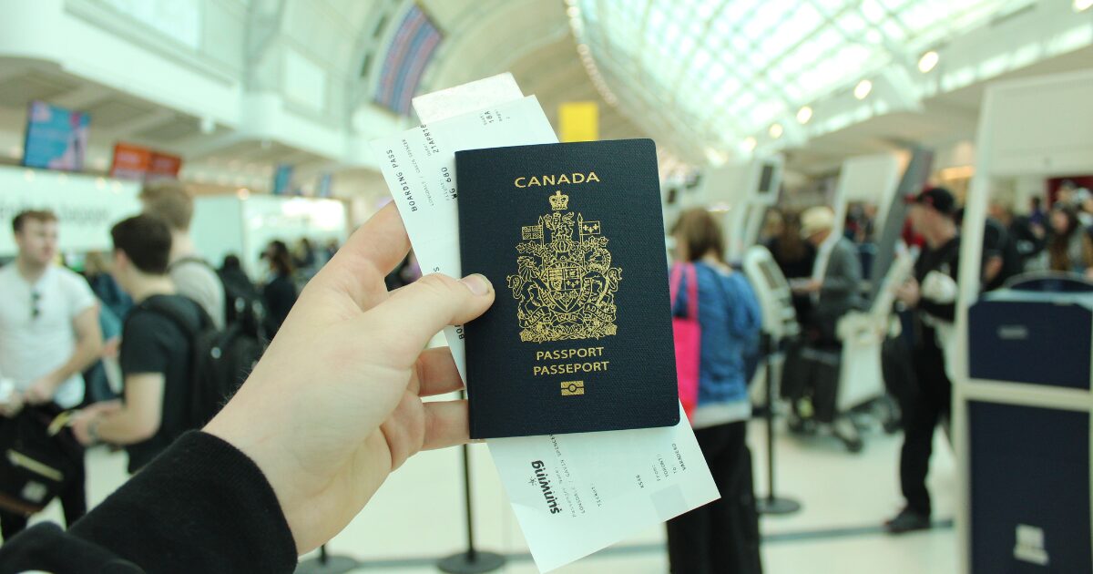 A Caucasian person's hand holding a Canadian passport in an airport with people in the background.