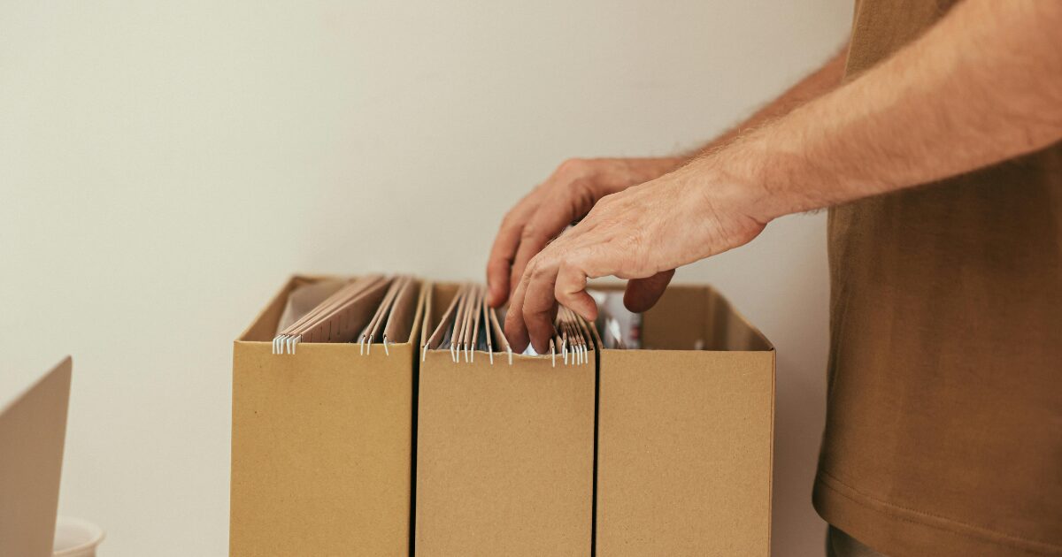 A close-up of a Caucasian man's hands sorting through file folders of documents in brown boxes.