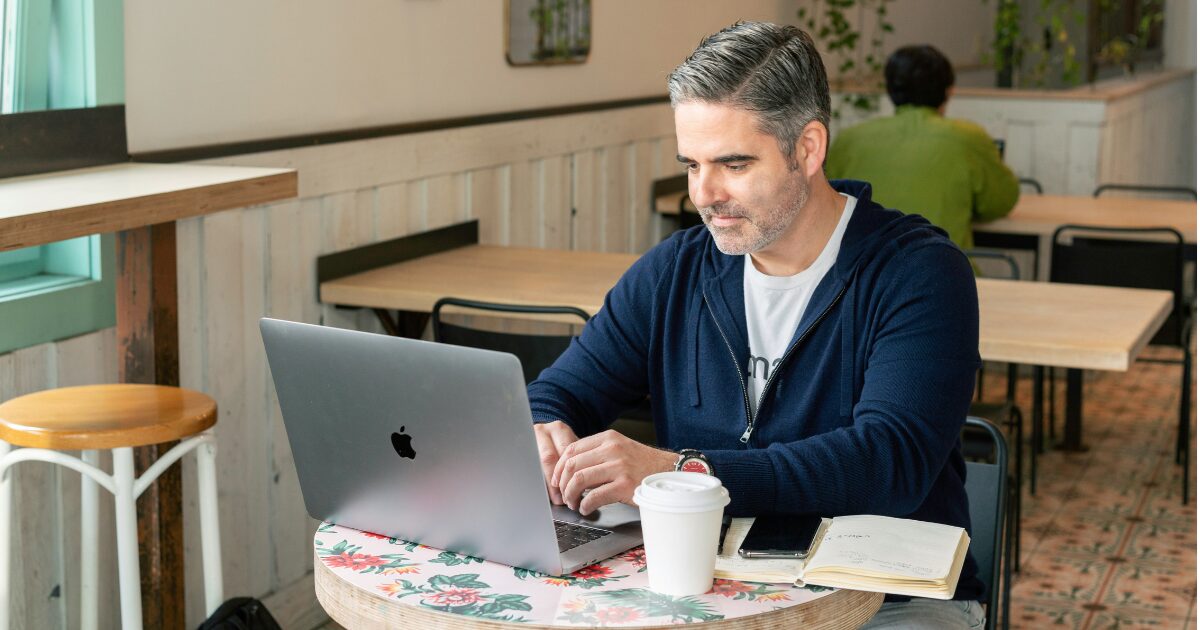 Middle-aged Caucasian man in a navy sweater typing on his laptop in a coffee shop.