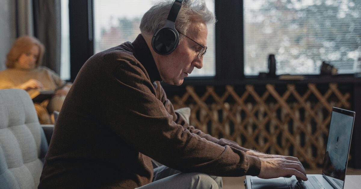 Elderly Caucasian man wearing a brown sweater, drafting a document, typing on his laptop.