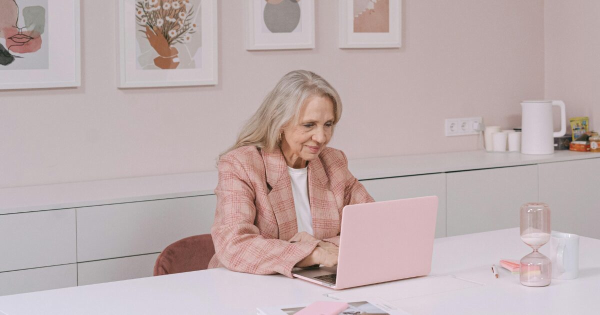 Elderly Caucasian woman wearing a pink blazer while she types on her laptop.