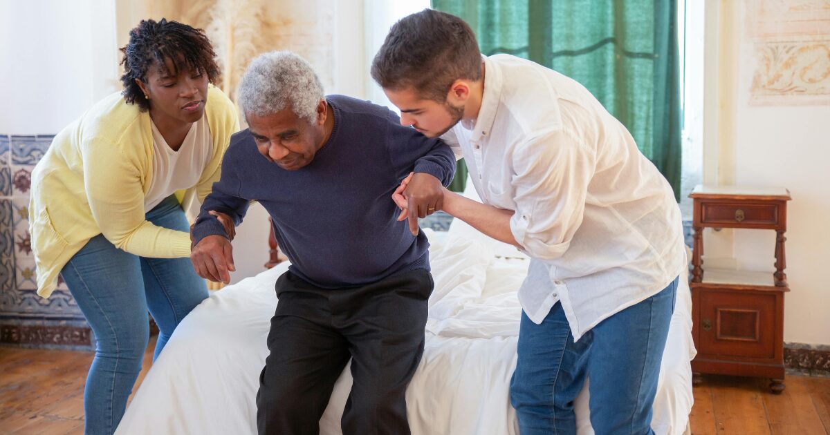 A Black adult woman and a young Caucasian man supporting an Elderly Black man to help him lay down in bed. 