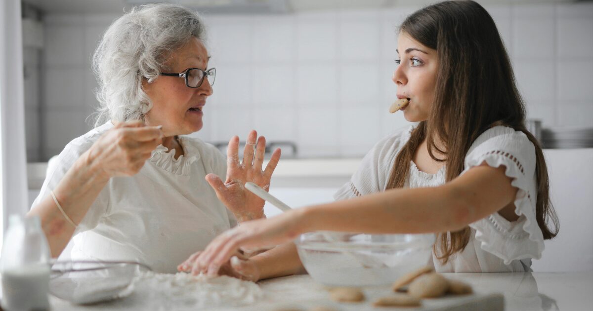 Elderly Caucasian woman making cookies with a young Caucasian woman. They are both wearing white and the young woman has a cookie in her mouth.