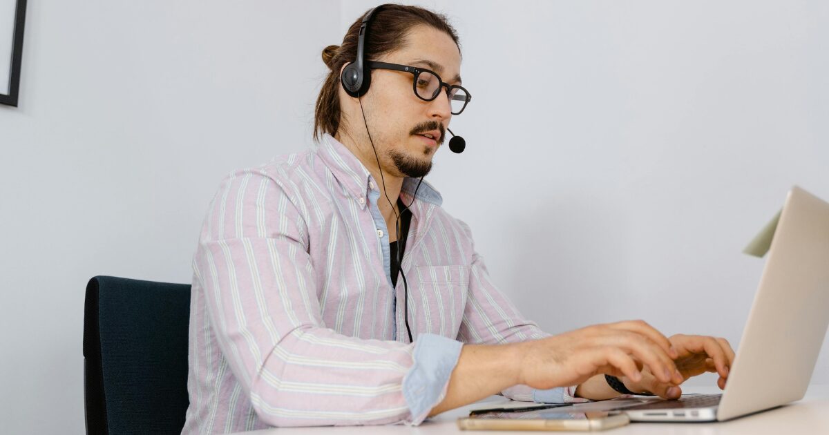 Young Caucasian man wearing eye glasses, a beige button-up shirt, and a headset, typing on a laptop.