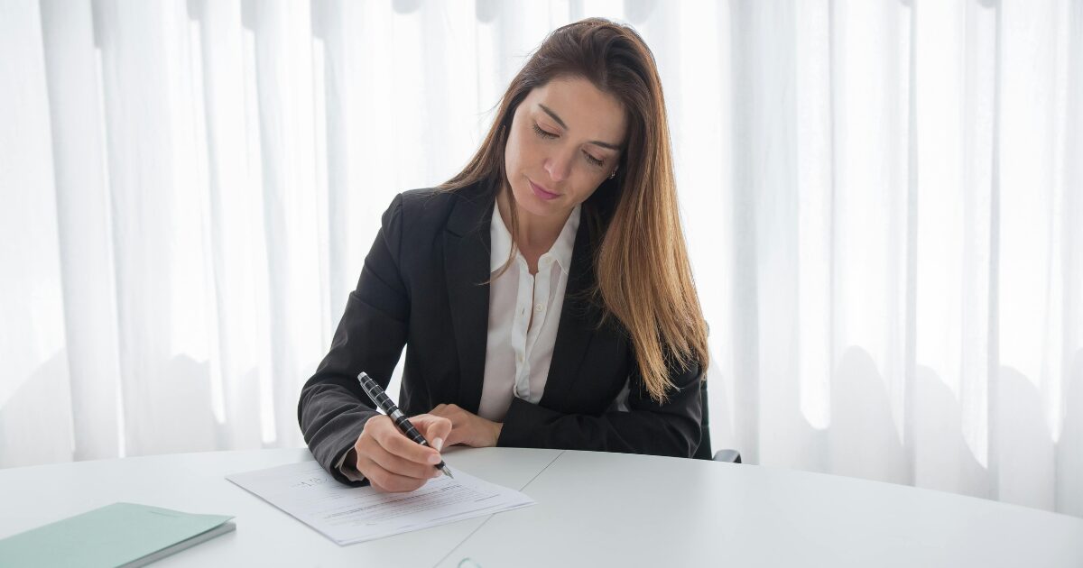 Young Caucasian woman with long brown hair, signing a document at a white desk. She is wearing a black blazer.