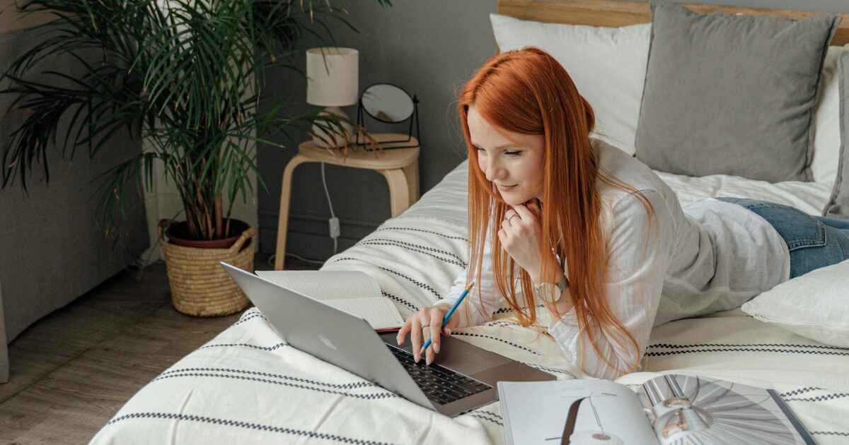 A young Caucasian woman laying on her bed as she works on her laptop.