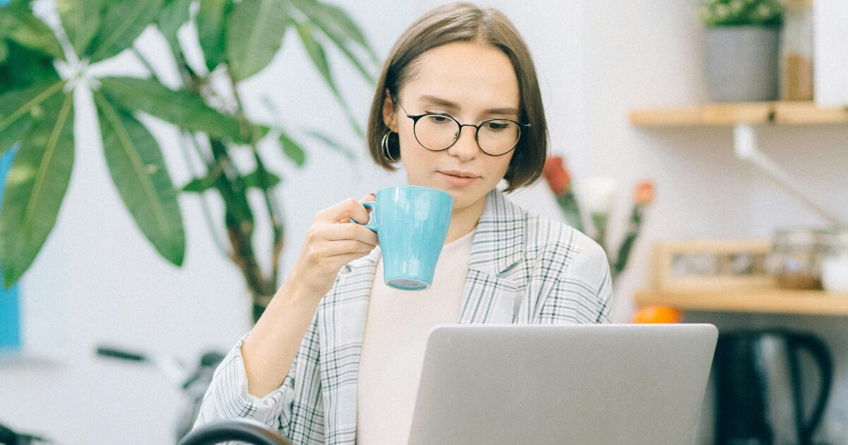 Young Caucasian woman working on her laptop with a turquoise mug in her hand. She has short brown hair and is wearing eye glasses.