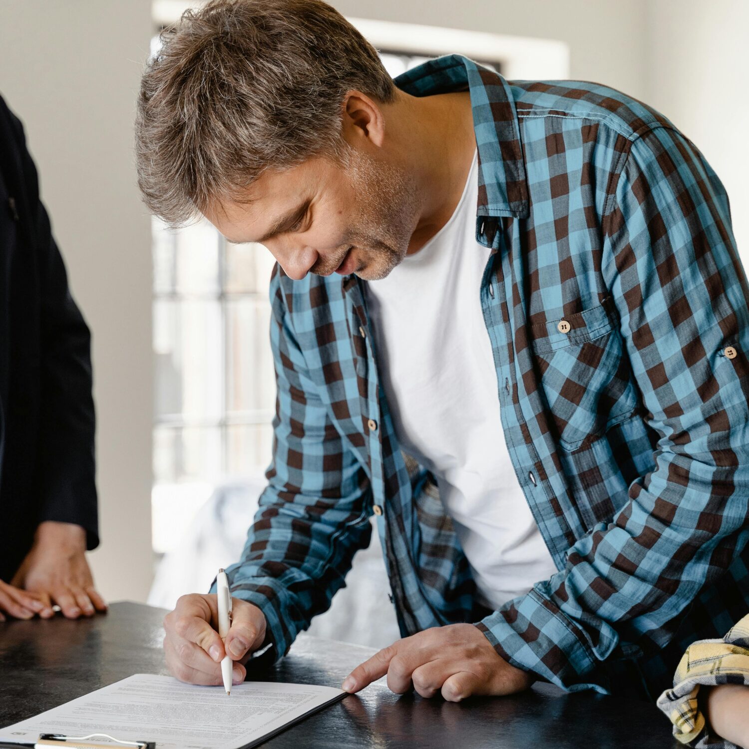 An older Caucasian man signing a document with a white pen. He is wearing a blue plaid button-up shirt with a white t-shirt underneath.